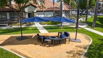 A blue umbrella shades a table and chairs in a sandy courtyard.